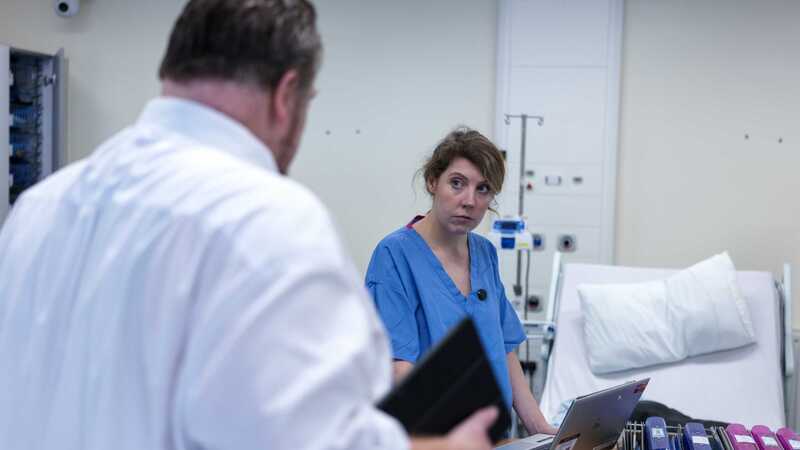 Photograph of a person in blue medical scrubs speaking to another in a white shirt within a hospital ward setting