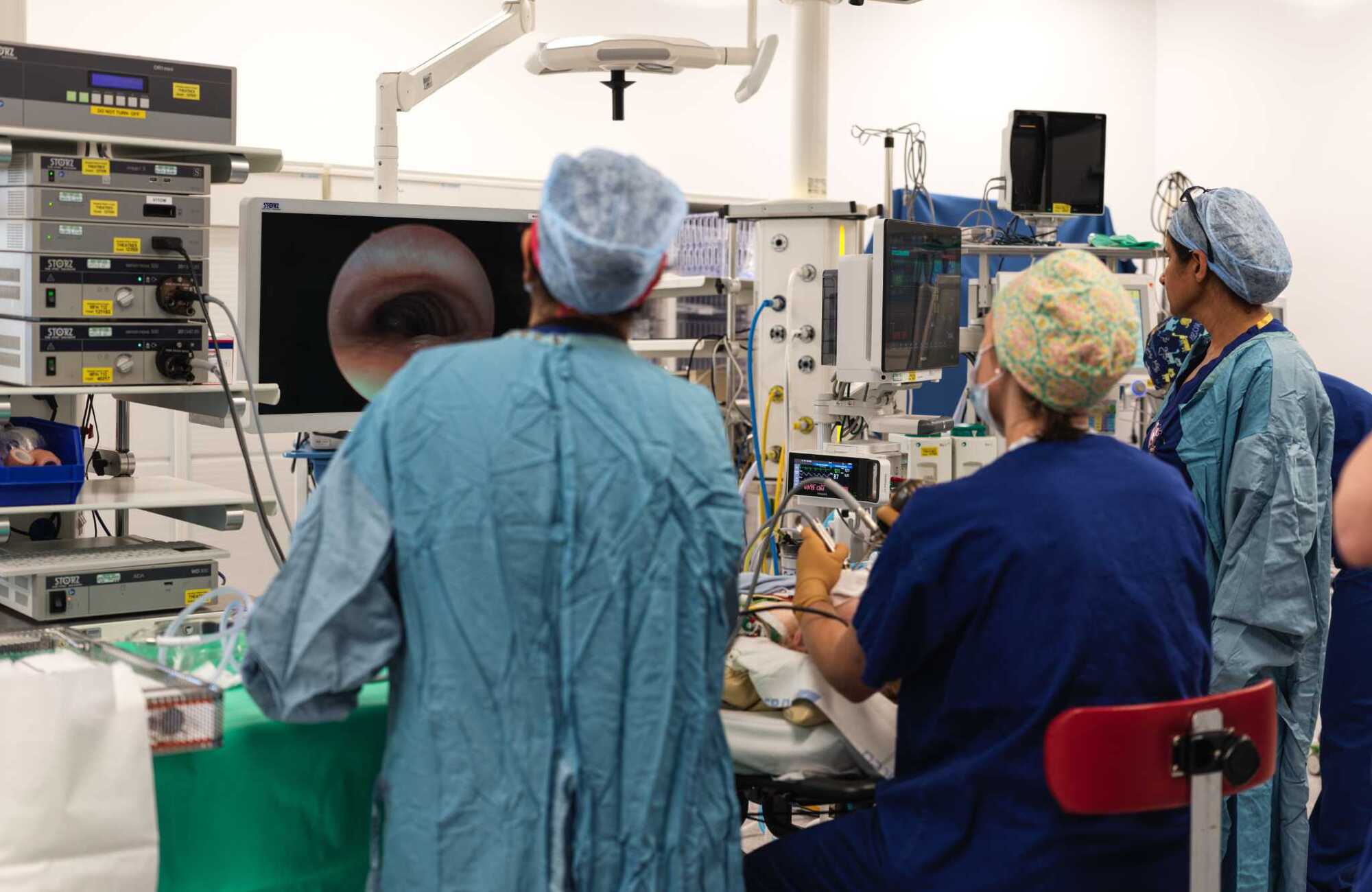 Photograph in an operating theatre of the backs of medics performing a procedure. They are viewing a screen in the background showing an image from the instruments they are holding.
