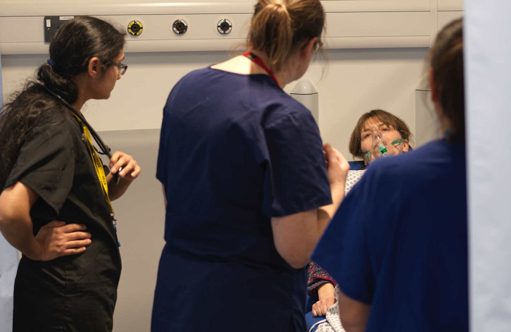 Photograph of a number of healthcare workers standing around a woman in a hospital bed with an oxygen mask over her face.