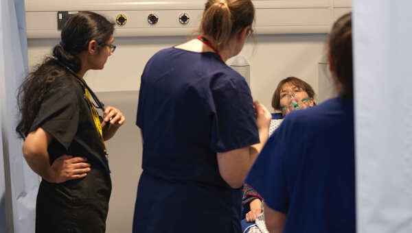 Photograph of a number of healthcare workers standing around a woman in a hospital bed with an oxygen mask over her face.